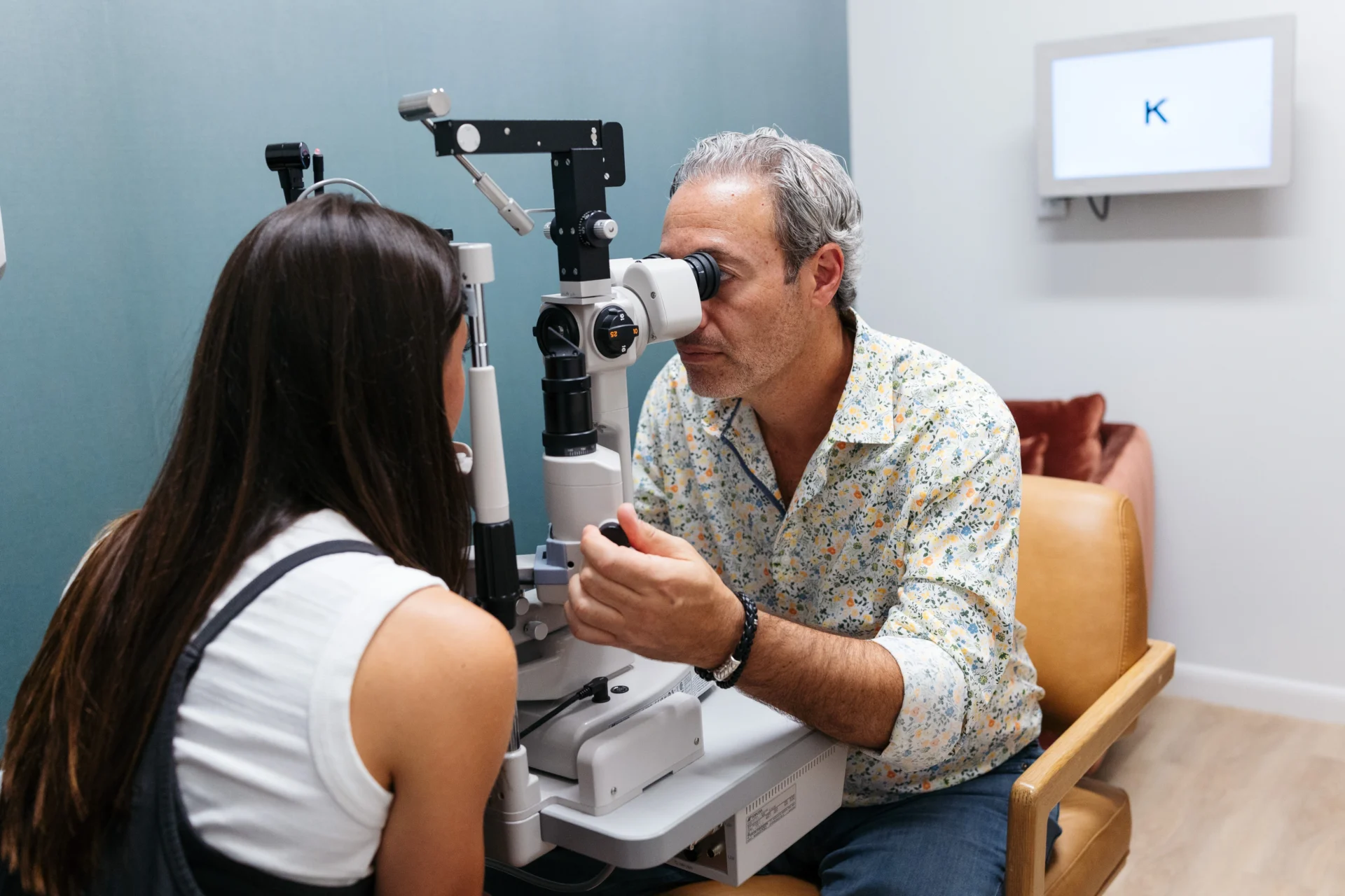 An optometrist examines a patient's eyes using a slit lamp in a modern eye clinic, with a monitor displaying the letter "K" in the background.