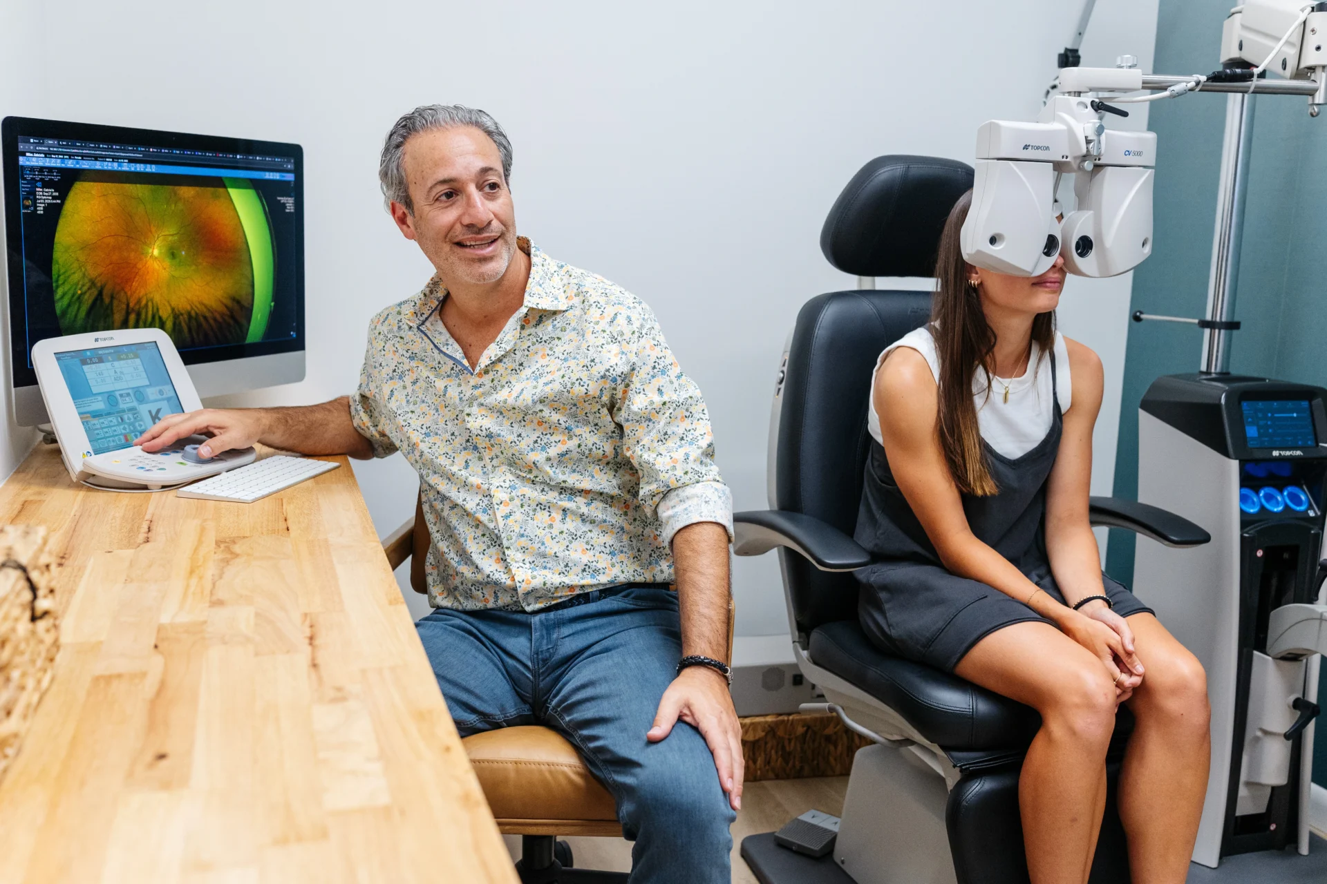 An eye doctor conducts a vision test for a patient using a phoropter, while examining retinal images on a computer screen in a clinic.