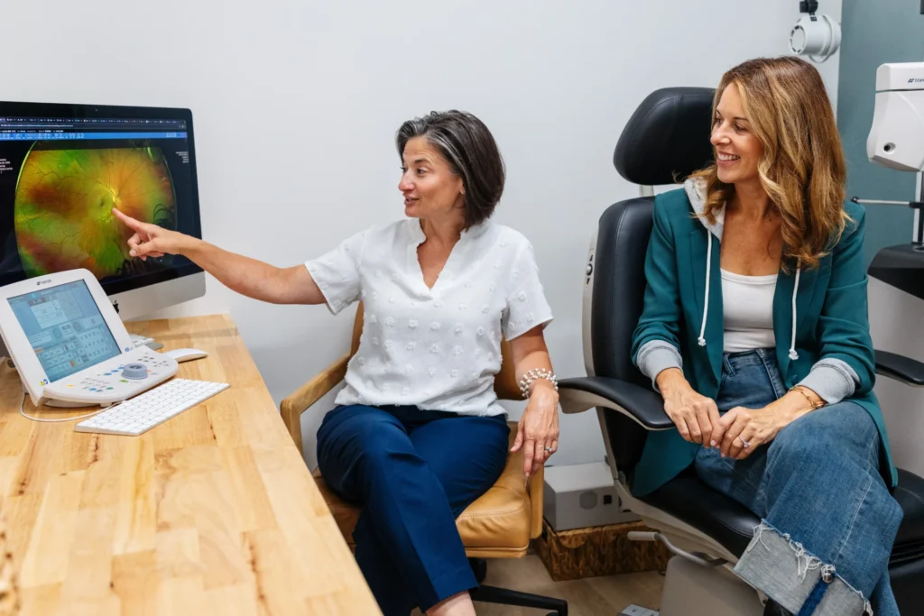 An eye care professional points at a retinal image on a computer screen while explaining it to a seated patient in an exam room.