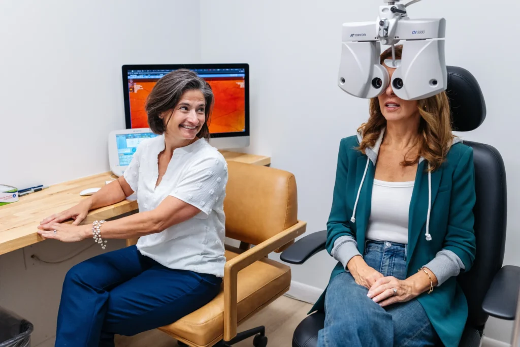 An eye doctor conducts a vision test on a seated patient using a phoropter, with retinal images displayed on a computer monitor in the background.