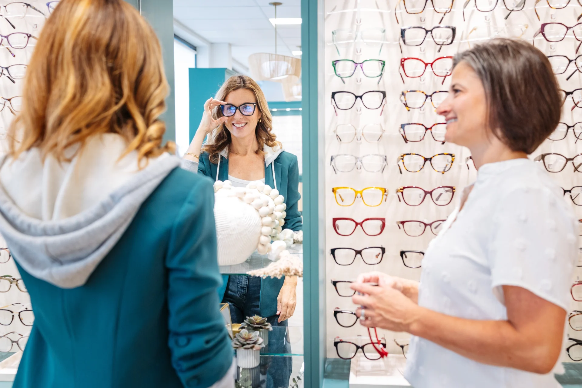 Two women stand in an eyewear store; one tries on glasses while looking in a mirror, and the other stands nearby holding a pair of red frames.