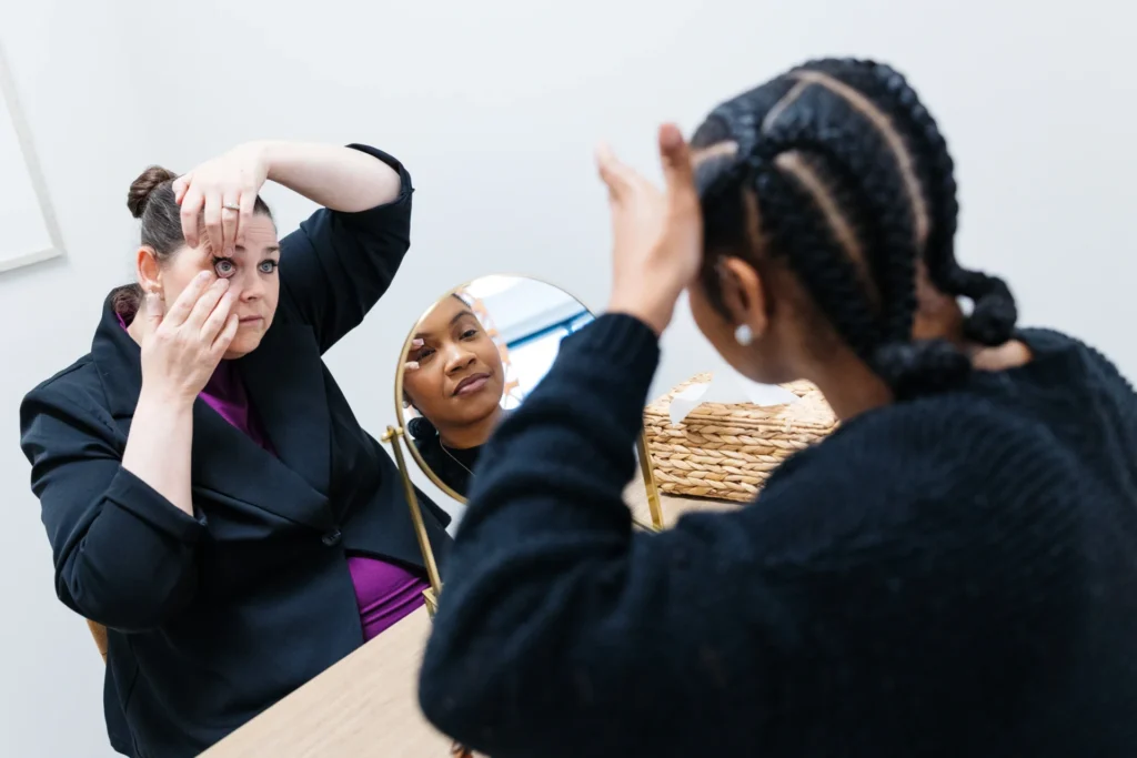 Two women sit at a table, one adjusting her eye makeup in a mirror, while the other touches her hair and looks at her own reflection.