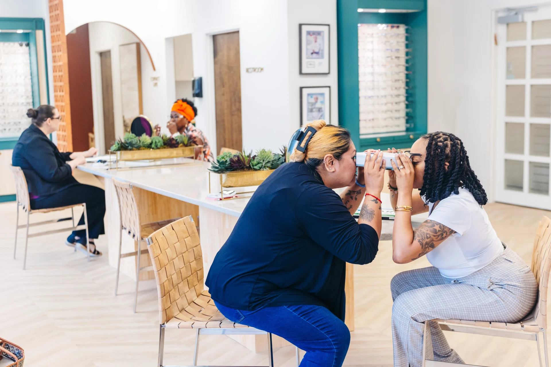 Two women sit facing each other, holding up optical devices to their eyes in an eyewear store, while two other people sit at a counter in the background.