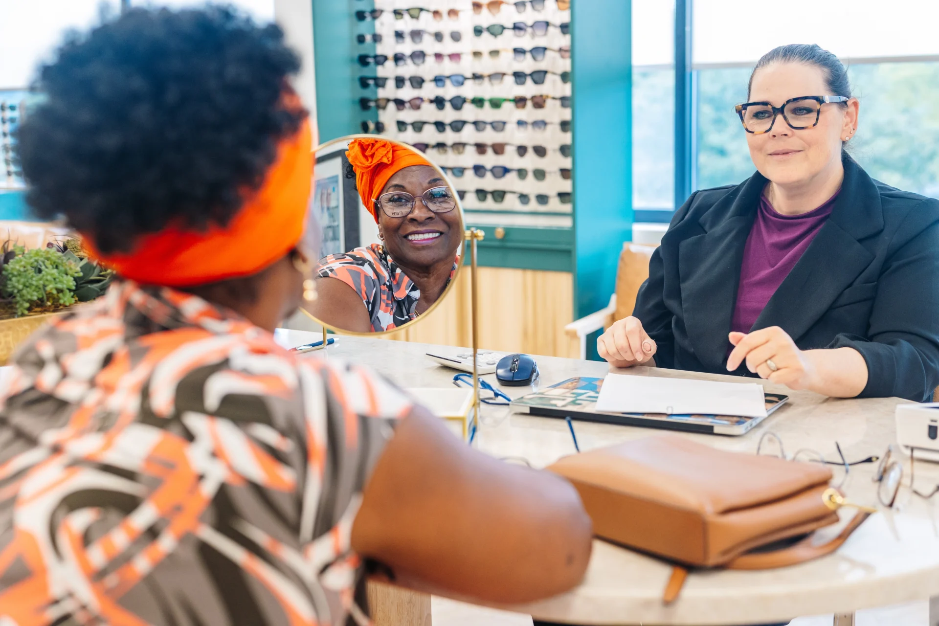 Two women sit across a table in an eyewear store, with one woman’s reflection visible in a mirror and sunglasses displayed in the background.