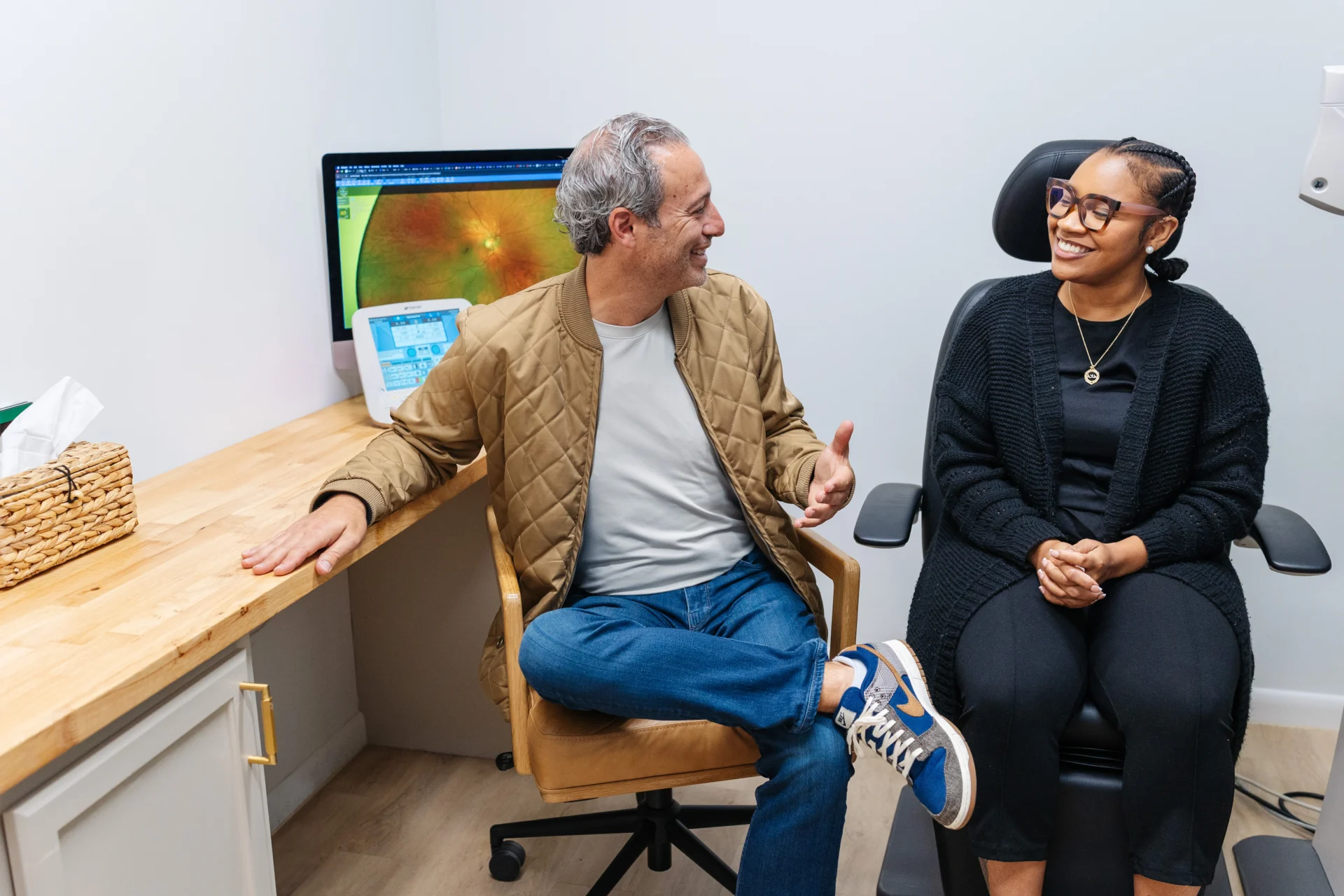 A man and a woman sit in an exam room, smiling and talking; a retinal image is displayed on a computer screen in the background.