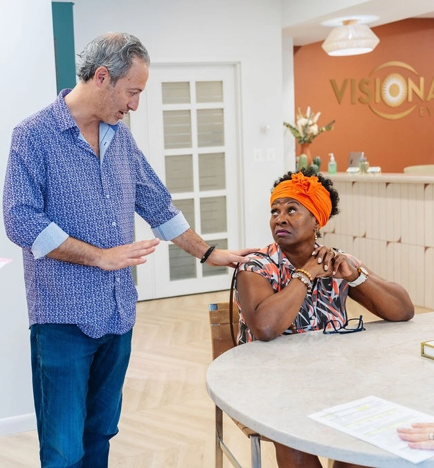 A man stands next to a seated woman with an orange headwrap, resting his hand on her shoulder as they talk in a modern office setting.