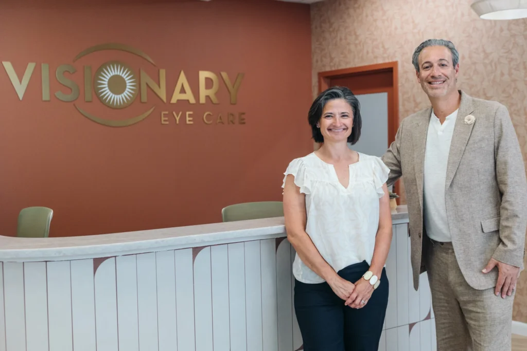 visionary-headshots-1001 - Visionary Eye Care Two people stand smiling in front of the reception desk at Visionary Eye Care, with the business name visible on the wall behind them.
