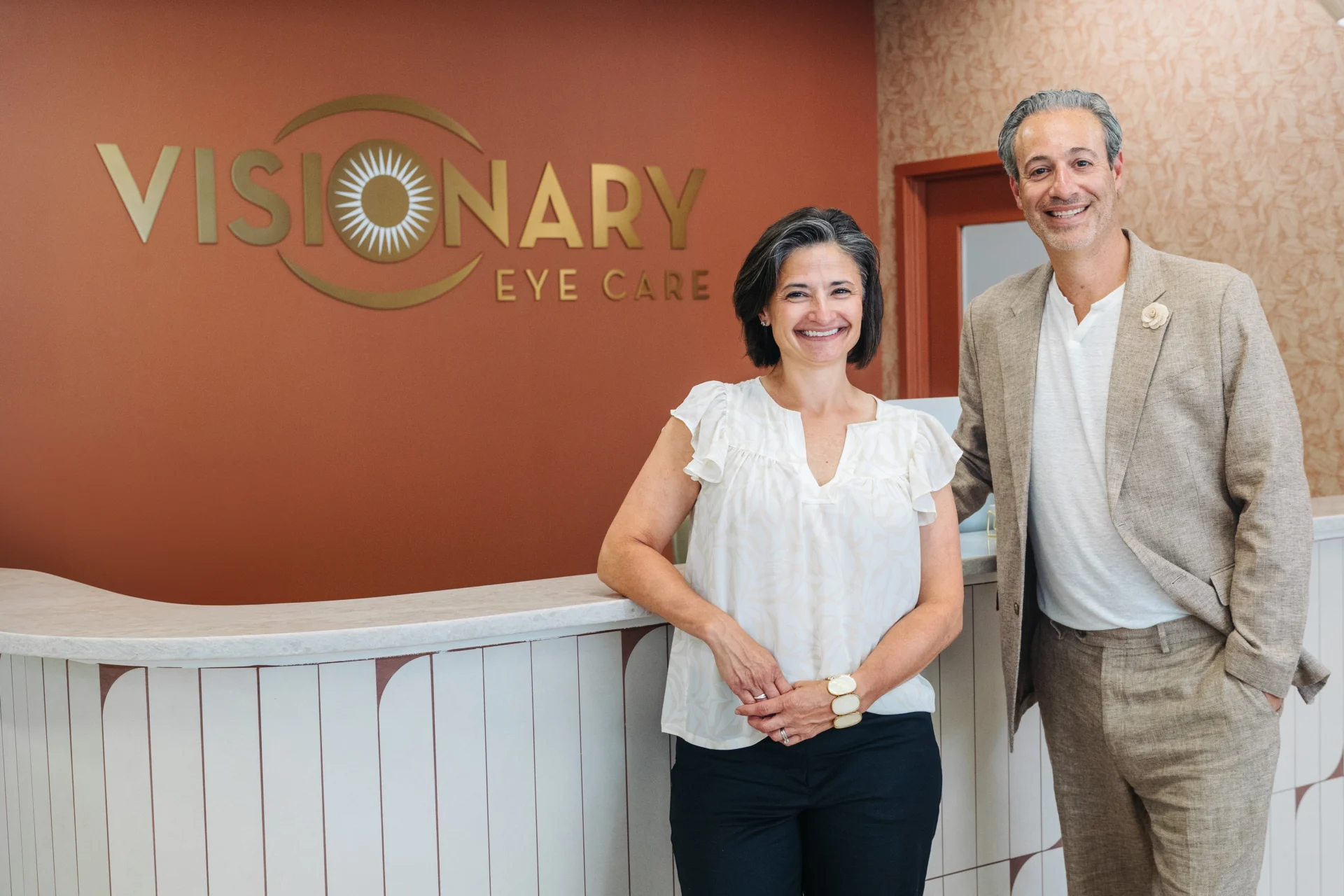 Two people stand smiling at the reception desk of Visionary Eye Care, with the clinic’s logo visible on the wall behind them.