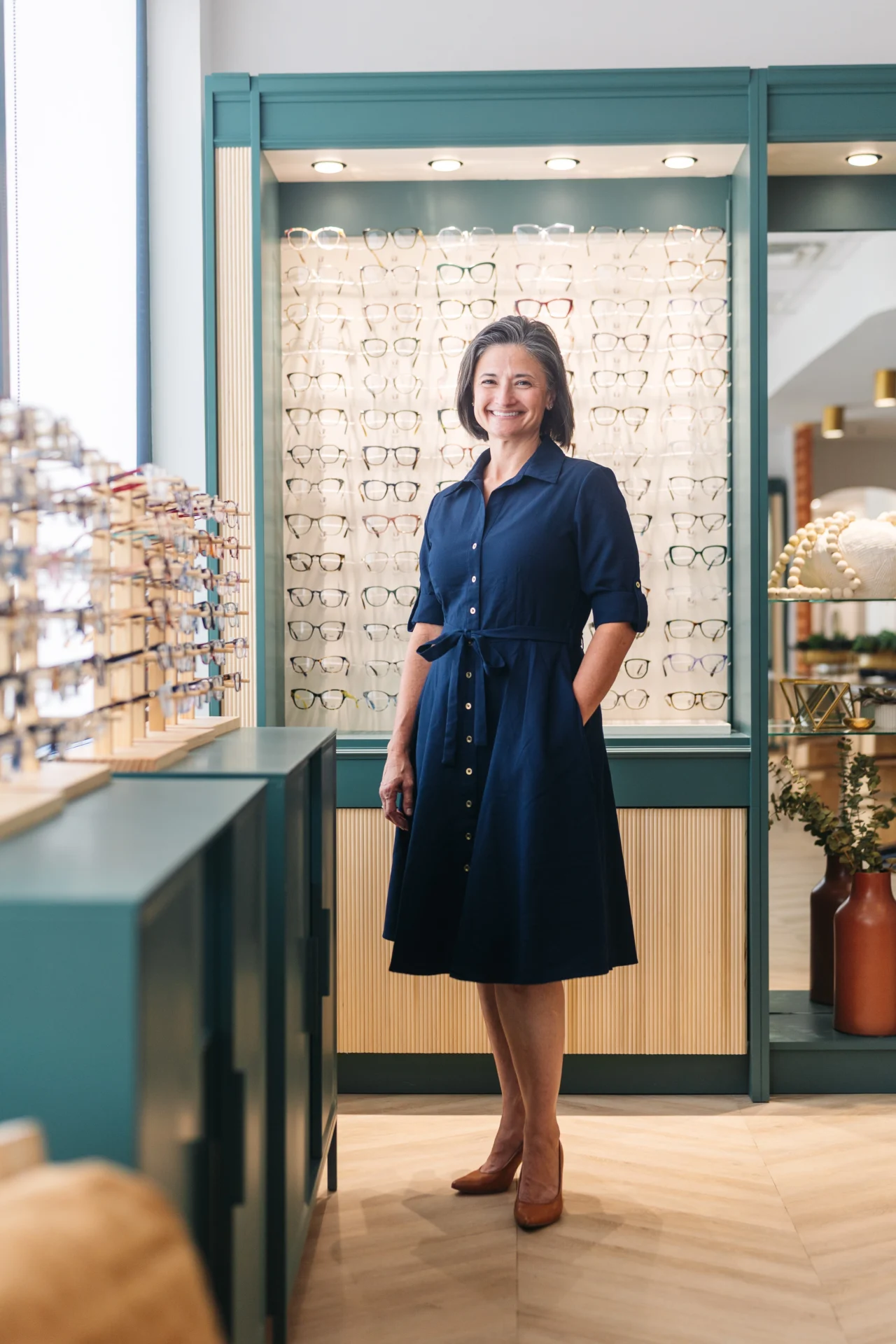 A woman wearing a navy blue dress stands in an eyewear store, surrounded by display racks of eyeglasses.