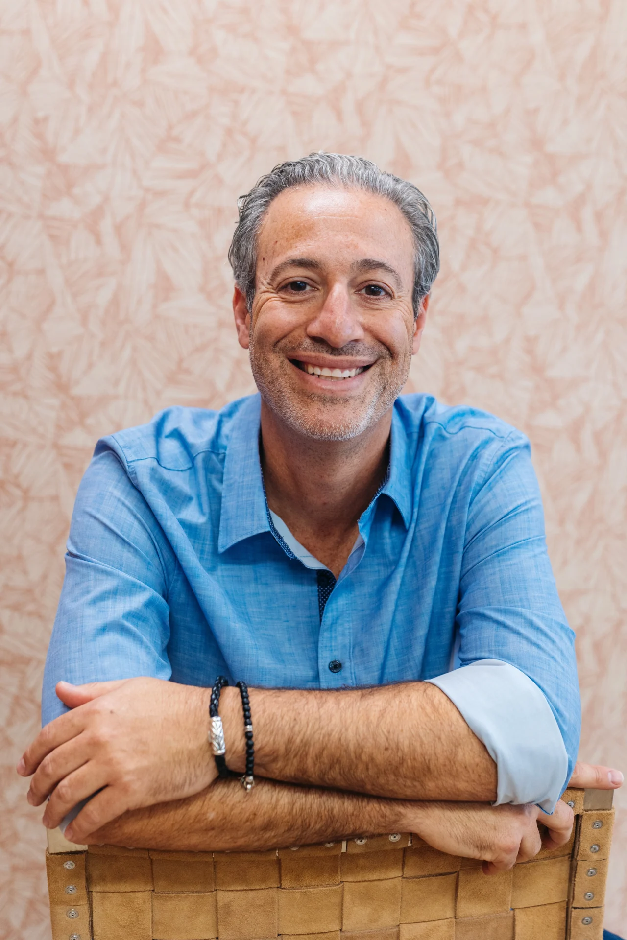 A man with gray hair in a blue shirt sits on a chair, smiling at the camera with his arms resting on the chair back. The background features a light, patterned wall.