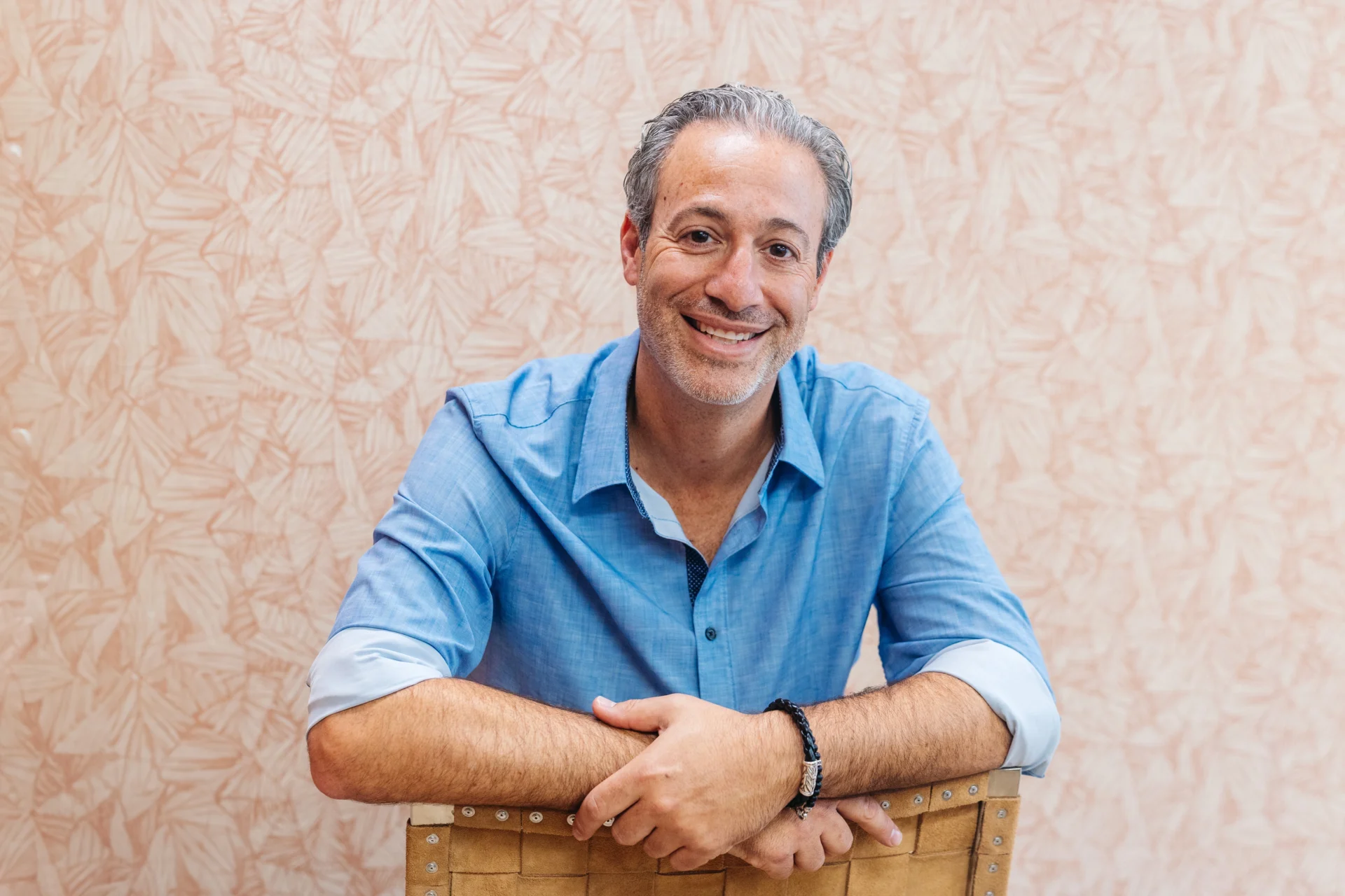 A man with short gray hair and a trimmed beard, wearing a light blue shirt, sits on a chair and smiles at the camera against a patterned beige background.