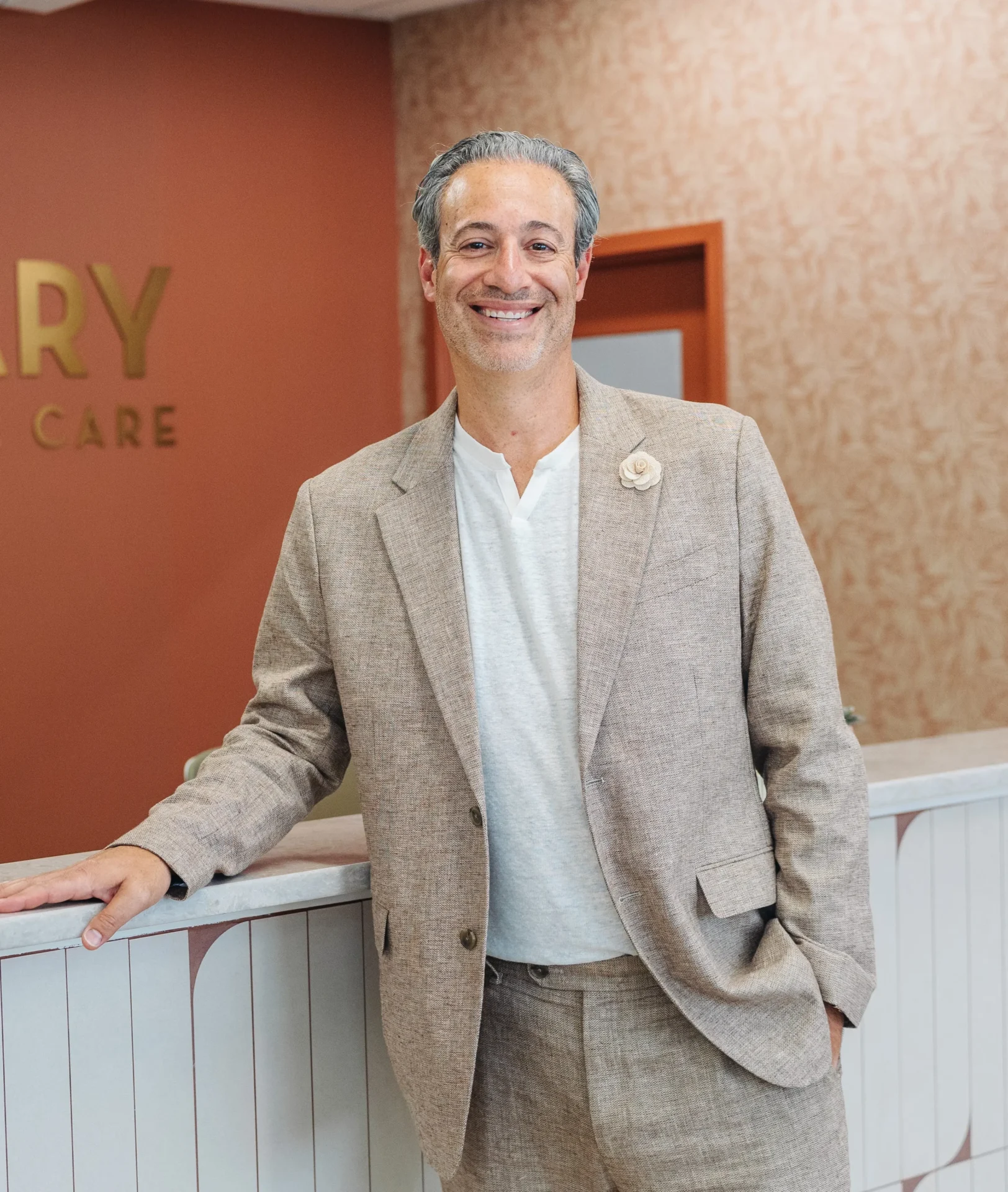A man in a beige suit and white shirt stands smiling by a reception counter in an office with pinkish walls and gold lettering in the background.