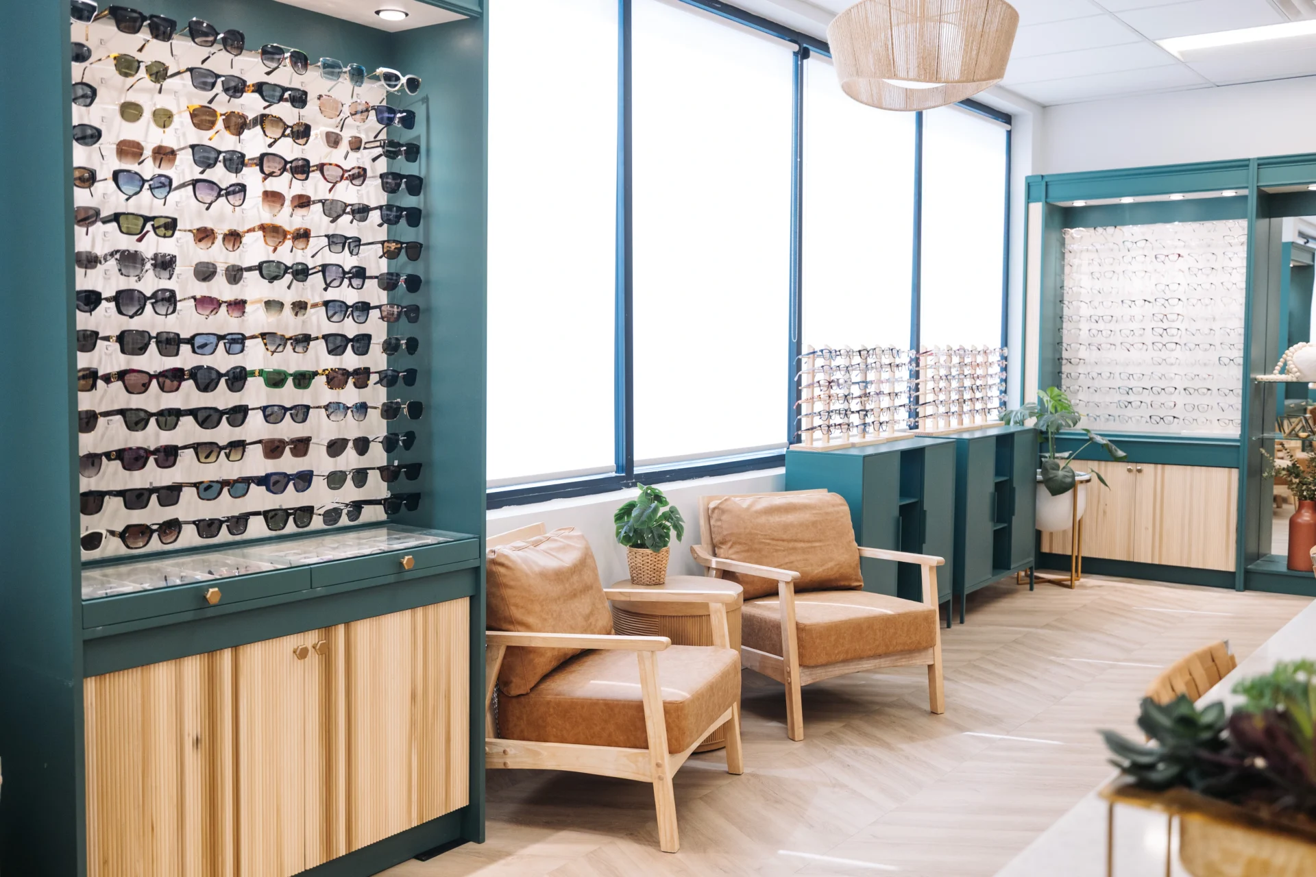 A modern eyewear store with display racks of sunglasses and eyeglasses, two tan chairs, and large windows letting in natural light.