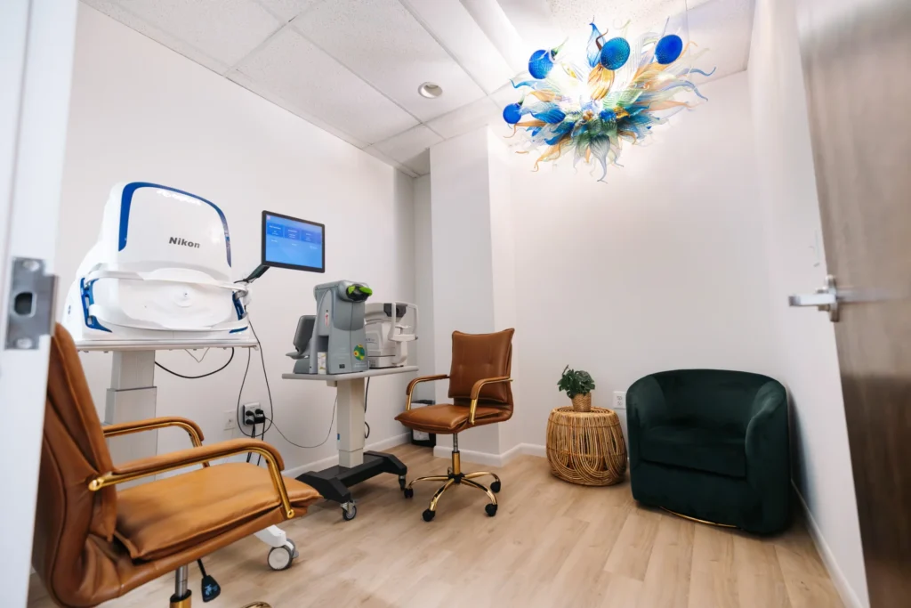 A small, modern eye exam room with ophthalmic equipment, two brown chairs, a green armchair, a side table with a plant, and a decorative blue ceiling light fixture.