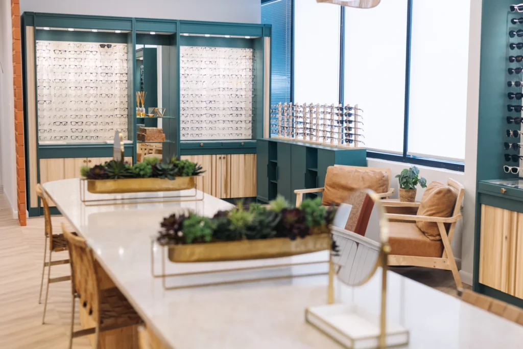 Modern optometry office with display shelves of eyeglasses, seating area with chairs, large windows, and decorative plants on a counter.