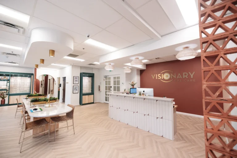 visionary-interiors-1078 - Visionary Eye Care Modern eye care clinic reception area with a front desk, computer, decorative lighting, seating area, and "Visionary Eye Care" sign on a rust-colored accent wall.