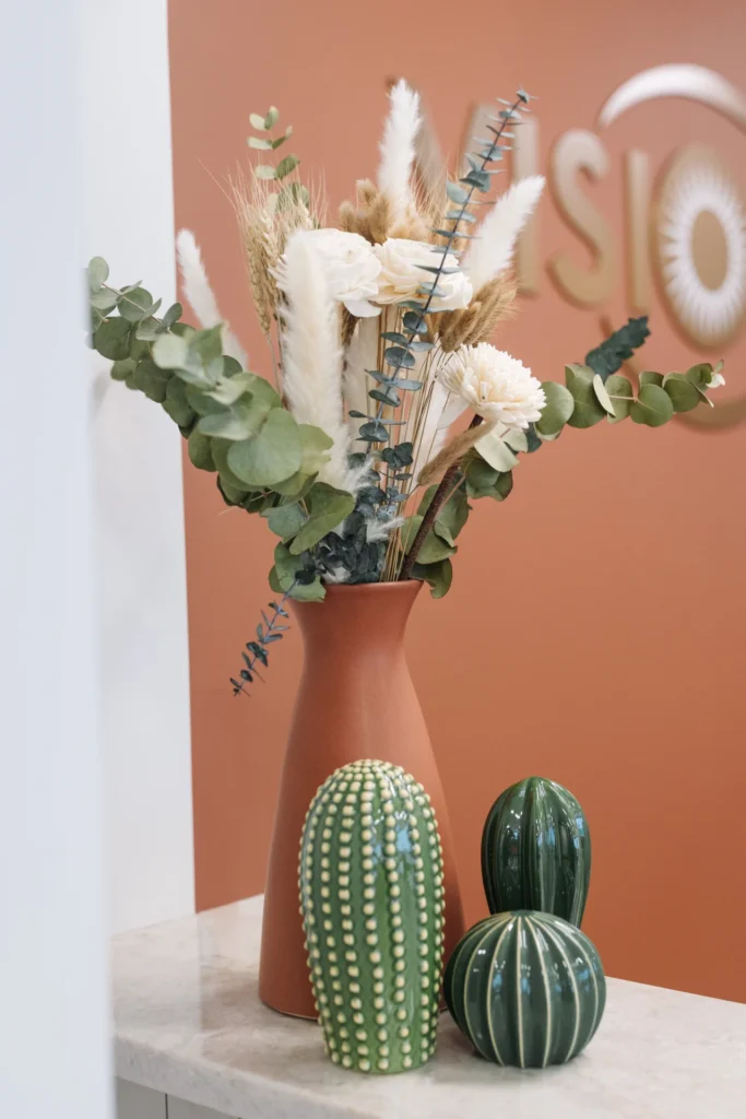 A terracotta vase with dried flowers and greenery sits on a marble surface beside three decorative ceramic cacti, against a rust-colored wall with partial signage.