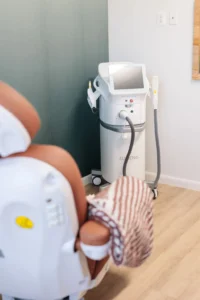 A medical device with a screen and two handheld attachments stands near a wall; a treatment chair with a striped blanket is visible in the foreground.