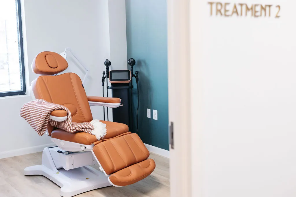 A modern treatment room with an empty brown reclining chair, a striped blanket, and medical equipment, viewed through a doorway labeled "TREATMENT 2.