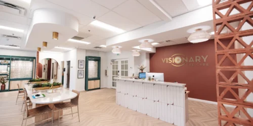 Modern eye care clinic reception area with a front desk, computer, decorative lighting, seating area, and "Visionary Eye Care" sign on a rust-colored accent wall.